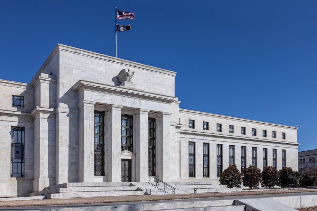 Front view of a white marble government building with tall columns, an eagle relief above the entrance, wide stairs, and flags against a blue sky.