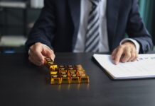 The price of gold continues to rise, up 11 percent from this year’s low Businessperson in a dark suit places gold bars on a desk beside a document.